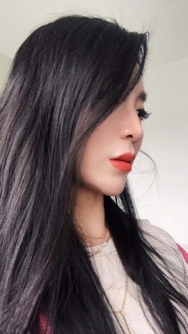 A young oriental female, with long dark straight hair. Near side of her face/eye partially cloaked by her hair. Wearing a light coloured sweater, plain background.